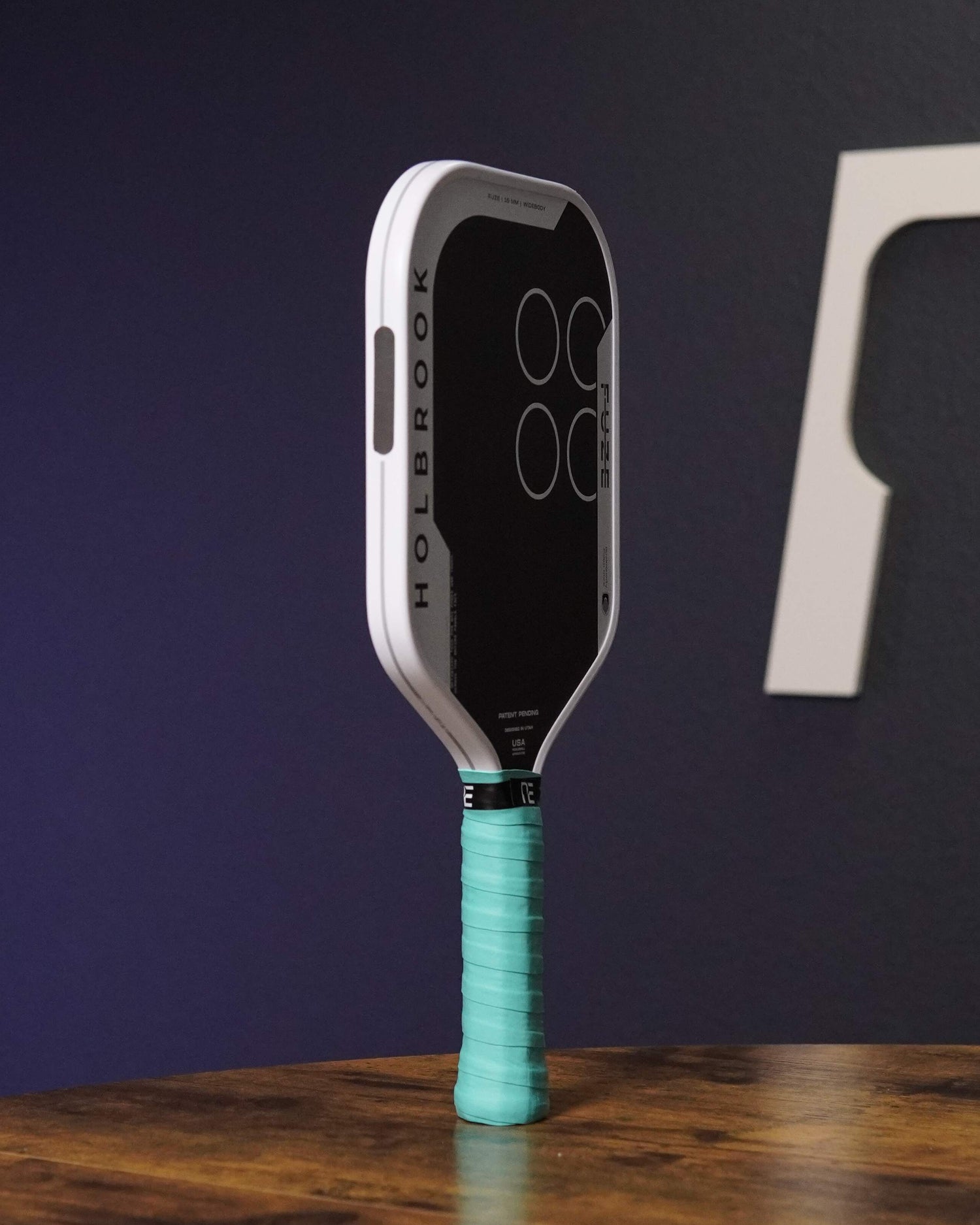 Pickleball paddle with teal handle on a wooden surface against a dark background with a logo.